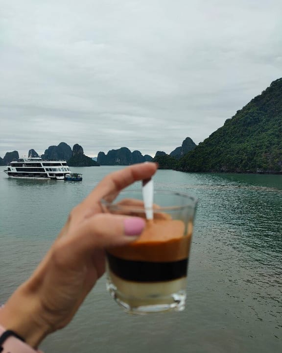 A glass of Vietnamese egg coffee held up against the karsts of the bay